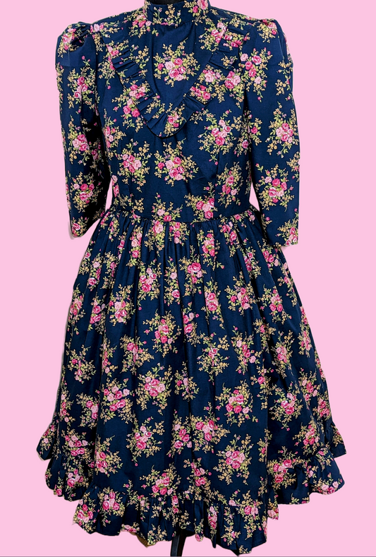 This Blue Floral Prairie Dress features beautiful pink flowers on a navy cotton fabric, with a 1970s-style vintage dress and Edwardian-inspired bodice. A timeless and elegant tea-length dress reminiscent of the iconic Little House on the Prairie