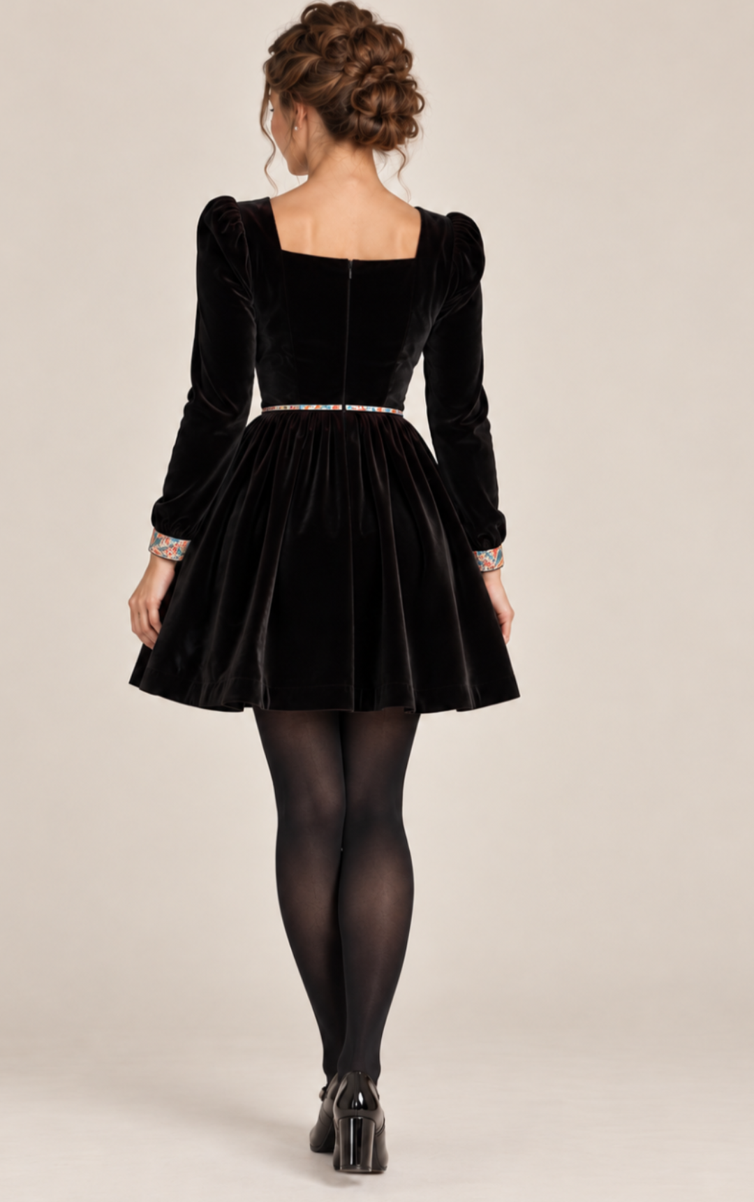 A woman shown from the back wearing a black velvet long-sleeve dress with a square neckline, puffed sleeves, a flared skirt, and a thin multicolor beaded waistband, paired with black tights and black heels against a plain beige studio background.