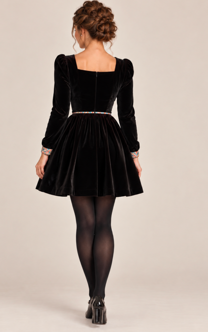 A woman shown from the back wearing a black velvet long-sleeve dress with a square neckline, puffed sleeves, a flared skirt, and a thin multicolor beaded waistband, paired with black tights and black heels against a plain beige studio background.